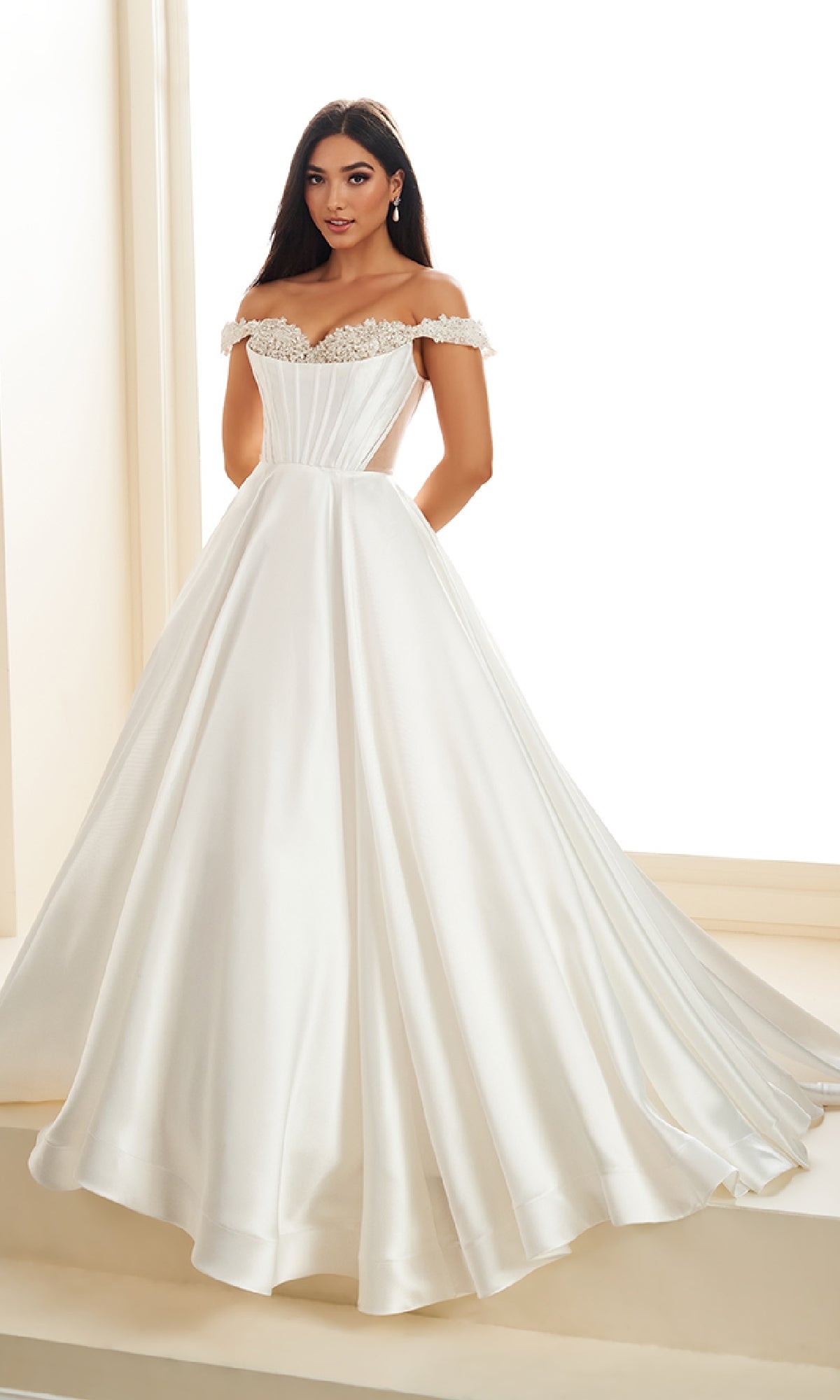 A woman stands indoors in front of large windows, wearing the Ellie Wilde EW36100 long prom dress—an off-the-shoulder ball gown featuring a corset bodice and a full, floor-length skirt.
